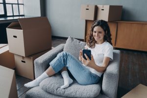 Happy apartment buyer among boxes. Young european woman posting news and smiling.